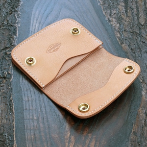 May include: A light tan leather card holder featuring white stitching, four brass snap fasteners, and a small embossed logo on the interior flap. The wallet is constructed with a smooth grain exterior and a natural suede interior texture.
