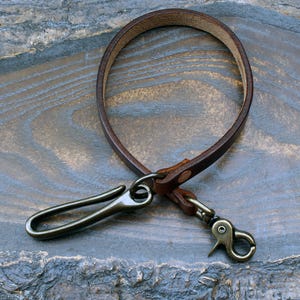 Könnte beinhalten: Ein brauner Leder-Schlüsselhalter mit Metallverschluss und Haken. Die Lederschlaufe ist mit einem Metallverschluss und Haken verbunden, um Schlüssel oder andere kleine Gegenstände zu halten. Der Schlüsselhalter besteht aus braunem Leder und Metall.