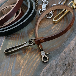 May include: A brown leather key holder with a brass clasp and swivel hook. The key holder is made of a thin strip of leather and has a loop design. Other leather strips and metal hardware are visible in the background.