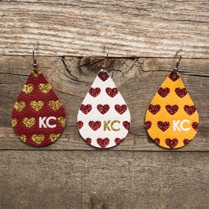May include: Three pairs of dangle earrings with a teardrop shape. Each earring is covered in glitter and features a pattern of red hearts. The earrings are red, white, and yellow. The text "KC" is printed on each earring.
