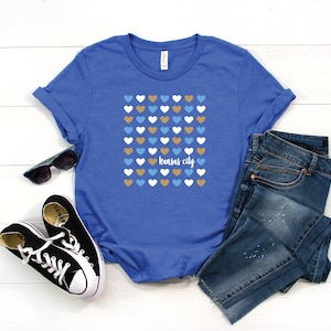 Puede incluir: Una camiseta azul real con un gr&aacute;fico de una cuadr&iacute;cula de corazones en azul, blanco y dorado. El texto "kansas city" est&aacute; impreso en blanco debajo de los corazones.