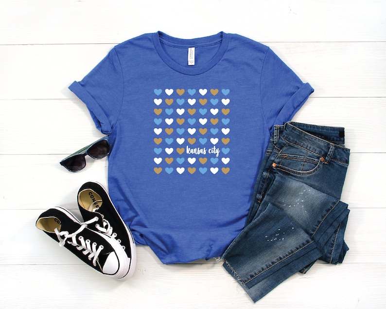 Puede incluir: Una camiseta azul real con un gr&aacute;fico de una cuadr&iacute;cula de corazones en azul, blanco y dorado. El texto "kansas city" est&aacute; impreso en blanco debajo de los corazones.