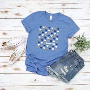 Puede incluir: Una camiseta azul con un patr&oacute;n de corazones dorados, blancos y azules. El texto "Kansas City" est&aacute; impreso en blanco en la camiseta.