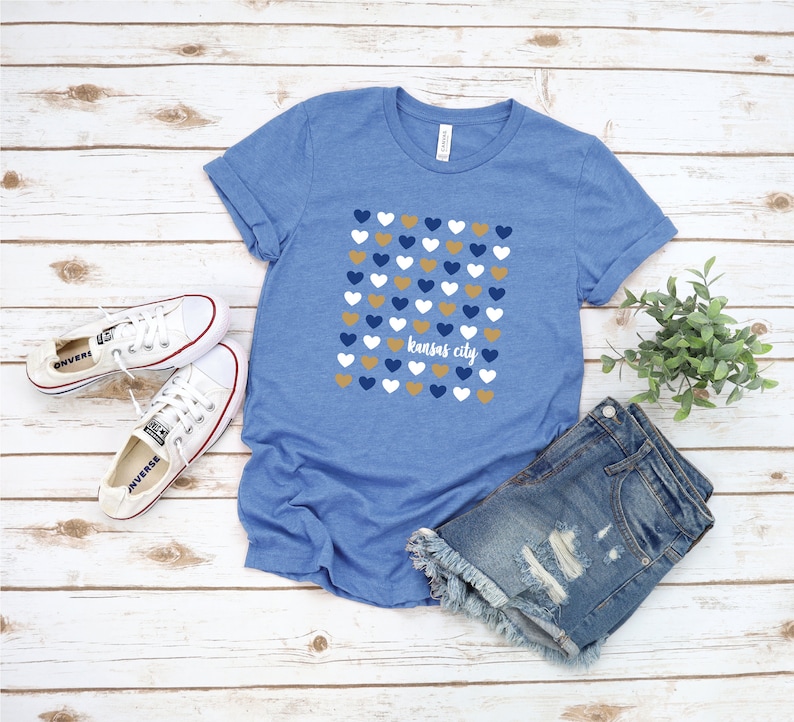 Puede incluir: Una camiseta azul con un patr&oacute;n de corazones dorados, blancos y azules. El texto "Kansas City" est&aacute; impreso en blanco en la camiseta.