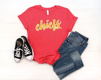 May include: A red t-shirt with the word "chiefs" in yellow lettering and a small heart above the word. The shirt is laying on a white wooden surface with a pair of black sneakers and blue jeans.