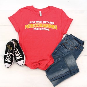 May include: Red t-shirt with the text "I just want to thank Patrick Mahomes for existing." in white and yellow letters. The shirt is paired with blue jeans and black sneakers.