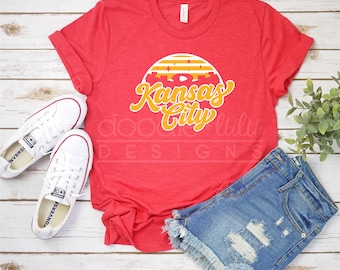 May include: Red t-shirt with a yellow and white graphic that says "Kansas City" with a sunset design behind the text.
