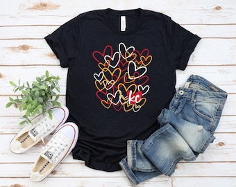 May include: A navy blue t-shirt with a graphic of white, red, and yellow hearts with the letters "KC" in the center. The shirt is laid out on a white wooden surface with a pair of white sneakers and blue jeans.