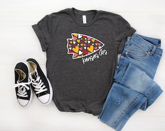 May include: Dark gray t-shirt with a graphic of an arrowhead filled with red, yellow, and white hearts, with the text "Kansas City" in a script font. The shirt is paired with blue jeans and black sneakers.