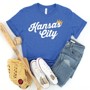 May include: A royal blue t-shirt with the text "Kansas City" in white with a gold crown above the word "Kansas". The shirt is laid out on a white surface with a baseball bat, a baseball, a baseball glove, and a pair of white sneakers.