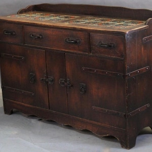 May include: A dark brown wooden cabinet with three drawers and two doors. The top surface features decorative tiles. The cabinet has metal hardware and is supported by four short legs. The wood has a distressed finish.