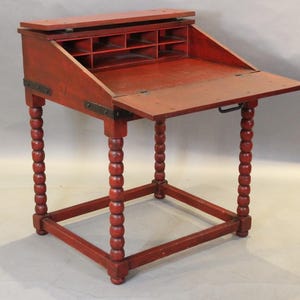 May include: A red wooden writing desk with a drop-down front and a shelf for storage. The desk has turned legs and is made of solid wood.