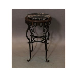 May include: A tall, round plant stand featuring a black and white marble top set within a dark metal frame. The base is constructed from wrought iron with curved, scroll-like legs and a circular support ring connecting the four legs near the bottom.