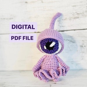 Puede incluir: Un juguete amigurumi de crochet con forma de extraterrestre morado con un solo ojo grande. El juguete está sentado sobre una superficie de madera blanca. El texto "DIGITAL PDF FILE" es visible en la imagen.