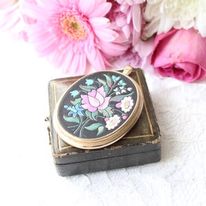 May include: An oval gold-tone locket featuring a black enamel inlay with a hand-painted floral motif. The design displays a central pink rose surrounded by smaller blue, white, and green flowers and leaves. The locket has a smooth metal border.