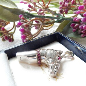 May include: A sterling silver horse head pendant with ruby accents. The pendant features a detailed horse head design with ruby eyes and a ruby-studded collar. The word "STERLING" is engraved on the side. The pendant is displayed in a blue box.