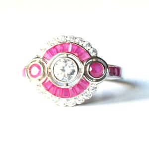 May include: A silver ring featuring a central clear gemstone encircled by a ring of pink gemstones. The ring has two smaller pink gemstones on either side of the central design, and is set with small clear gemstones around the pink ring.