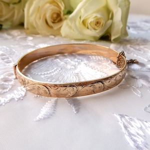 May include: A gold-coloured bangle bracelet with an engraved floral pattern. The hinged bracelet has a clasp and safety chain. The background features white roses and a white lace surface.