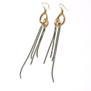 Puede incluir: Un par de pendientes dorados con un diseño de doble óvalo y cadenas colgantes de color plateado. Los pendientes tienen un cierre de gancho y están sobre un fondo blanco. Las cadenas miden unos 10 cm de largo.