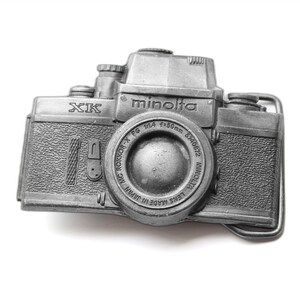 Könnte beinhalten: Eine silberfarbene Gürtelschnalle aus Metall in Form einer Minolta XK Kamera. Die Vorderseite zeigt den Schriftzug Minolta, XK und Objektivdetails wie MC ROKKOR-X PG 1:1.4 f=50mm. Die Schnalle hat eine strukturierte Oberfläche und eine rechteckige Öse.