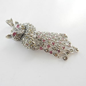 Peut inclure: Un pendentif en métal argenté en forme de paon, dont le corps et la queue sont incrustés de nombreuses petites pierres de marcassite gris foncé et agrémentés de petites pierres précieuses rondes rouge profond serties dans le métal.