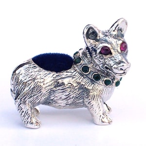 Puede incluir: Un cojín para alfileres plateado con forma de corgi con un cojín de terciopelo azul oscuro. El perro tiene ojos de gema roja y un collar de gema verde. Un artículo decorativo.