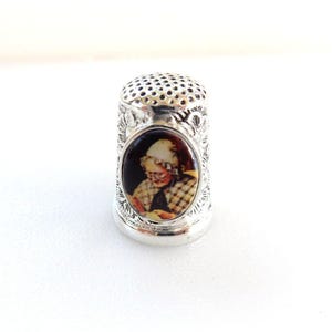 Könnte beinhalten: Ein silberner Fingerhut mit detailliertem floralem Design und einem kleinen ovalen Bild einer Person. Der Fingerhut hat eine perforierte Oberseite und einen glatten, abgerundeten Boden. Das Bild zeigt eine Person mit Hut und kariertem Hemd.