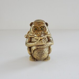 Op de afbeelding: Een goudkleurig apenbeeldje met een scharnierend deksel, dat een kleine bol vasthoudt. De aap heeft gedetailleerde gelaatstrekken, waaronder grote ogen en een contemplatieve uitdrukking. Het beeldje is waarschijnlijk van metaal en heeft een getextureerd oppervlak.