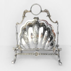 May include: A silver-plated biscuit barrel featuring a hinged shell-shaped lid with intricate floral and circular engravings. The container is supported by a decorative metal frame with a circular handle at the top and four curved feet at the base.