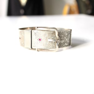 Puede incluir: Una pulsera de metal plateado diseñada para parecer un cinturón con hebilla. La superficie presenta una textura cepillada, patrones de estrellas grabados y pequeñas piedras rojas incrustadas. La pulsera tiene un panel central liso.