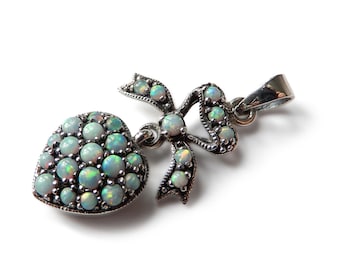 Op de afbeelding: Zilveren hanger met een hart- en strikdesign, bezet met opalescerende edelstenen. Het hart en de strik zijn bedekt met kleine, ronde edelstenen. De hanger heeft een oogje voor een ketting. De edelstenen hebben blauwe, groene en witte tinten.