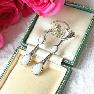 May include: A pair of drop earrings featuring two oval and teardrop-shaped opals set in silver metal. The earrings have intricate textured metal detailing at the top and between the stones, creating a long, dangling design with a polished finish.