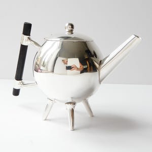 May include: A spherical silver teapot featuring a polished metallic finish, a long angled spout, and a lid with a small round knob. The teapot stands on three short silver legs and includes a vertical black handle attached with silver hardware.