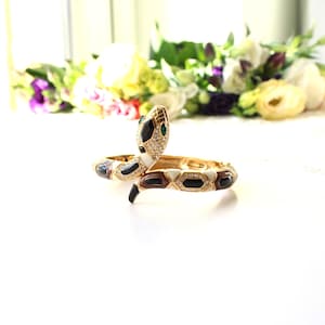 May include: A gold-tone hinged snake bracelet featuring a coiled design with black and white enamel segments, clear crystal accents, and small green crystal eyes. The metal body is textured with a scale-like pattern throughout the entire piece.