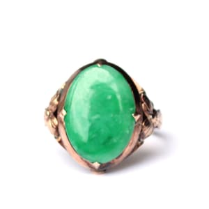 May include: A vintage ring with a large, oval, bright green jade stone set in a gold-toned metal band. The band has decorative floral accents on the sides. The ring is set against a white background. The jade stone is approximately 2.5 cm in length.