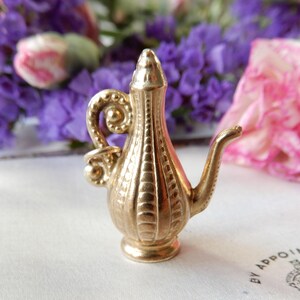 May include: A small, gold-coloured decorative jug with a curved handle and spout. The jug has a ribbed design and a pointed top. The background features purple and pink flowers.