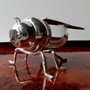 May include: A polished silver-toned metal insect-shaped container featuring a rounded head with antennae, a bulbous body, and six slender legs. The object has a reflective surface and a hinged lid that extends into a wing-like shape at the back.