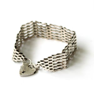 Puede incluir: Una pulsera plateada con un diseño tejido. La pulsera presenta un cierre de candado en forma de corazón. La pulsera de metal se muestra sobre un fondo blanco. La pulsera es una joya.