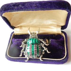 May include: A silver-toned beetle brooch with green and purple accents, set in a purple velvet jewelry box. The beetle has a green enamel shell with purple stripes and is adorned with small, sparkling stones. The box has a cream-colored satin lining.