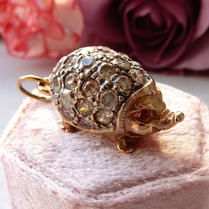 Puede incluir: Colgante de oro con forma de erizo, incrustado con numerosas piedras preciosas transparentes. El colgante tiene un pequeño lazo para una cadena y está sobre una superficie rosa, con flores rosas y moradas borrosas en el fondo.