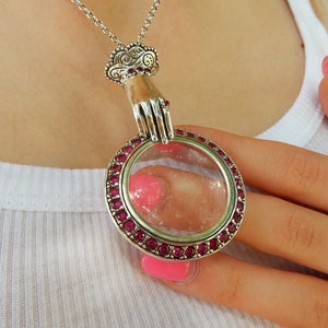 Op de afbeelding: Zilverkleurige hangerketting met een ronde, heldere glazen locket met een rand van kleine, rode edelstenen. De hanger wordt vastgehouden door een gedetailleerd handontwerp en hangt aan een fijne ketting. De ketting wordt gedragen over een witte top.