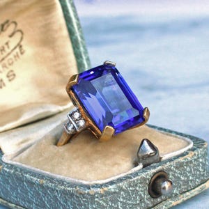 Op de afbeelding: Een vintage gouden ring met een grote, rechthoekige, diepblauwe edelsteen. De ring heeft een gouden band en accenten, met kleinere diamanten aan de zijkant. De ring wordt gepresenteerd in een vintage, lichtblauwe sieradendoos.
