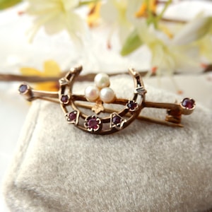 May include: A vintage gold-coloured brooch with a horseshoe design, three pearls, and small red gemstones. The brooch has a bar pin closure. The background is a soft, neutral colour.