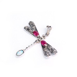 May include: A silver dragonfly pendant with ornate wing detailing. The pendant features three oval-cut, deep red gemstones and a single, oval, opalescent stone. The dragonfly's body and wings are intricately designed, creating a delicate and detailed piece of jewelry.