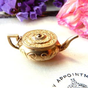 May include: A small, gold-colored teapot with intricate embossed detailing. The lid features a circular design, and the body has swirling patterns. The handle and spout are curved. The teapot is set against a backdrop of purple and pink flowers.