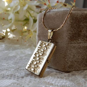 Puede incluir: Un collar con colgante rectangular dorado con un diseño texturizado. El colgante cuelga de una delicada cadena dorada. El collar se exhibe sobre una superficie suave de color neutro, con flores blancas en el fondo.