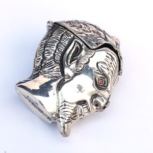 May include: A silver-colored, hinged box in the shape of a stylized animal head. The box features intricate detailing, including textured surfaces and a red gemstone eye. The lid is slightly ajar, revealing the interior.