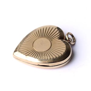 Puede incluir: Un relicario dorado en forma de corazón con un diseño de rayos de sol en la parte delantera. El relicario tiene un pequeño lazo en la parte superior para sujetar una cadena. El relicario está cerrado y parece ser de metal.