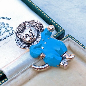 Könnte beinhalten: Vintage Katzenbrosche. Die Brosche zeigt einen silberfarbenen Katzenkopf und einen blau emaillierten Körper. Die Katze trägt ein blaues Outfit mit silbernen Akzenten. Die Brosche befindet sich in einer grün-weißen Schmuckschatulle.