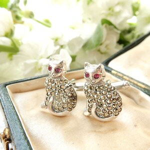 Puede incluir: Un par de gemelos de plata con diseño de gato. Los gatos están hechos de plata y tienen ojos rojos. Los gatos están cubiertos de pequeñas piedras brillantes.